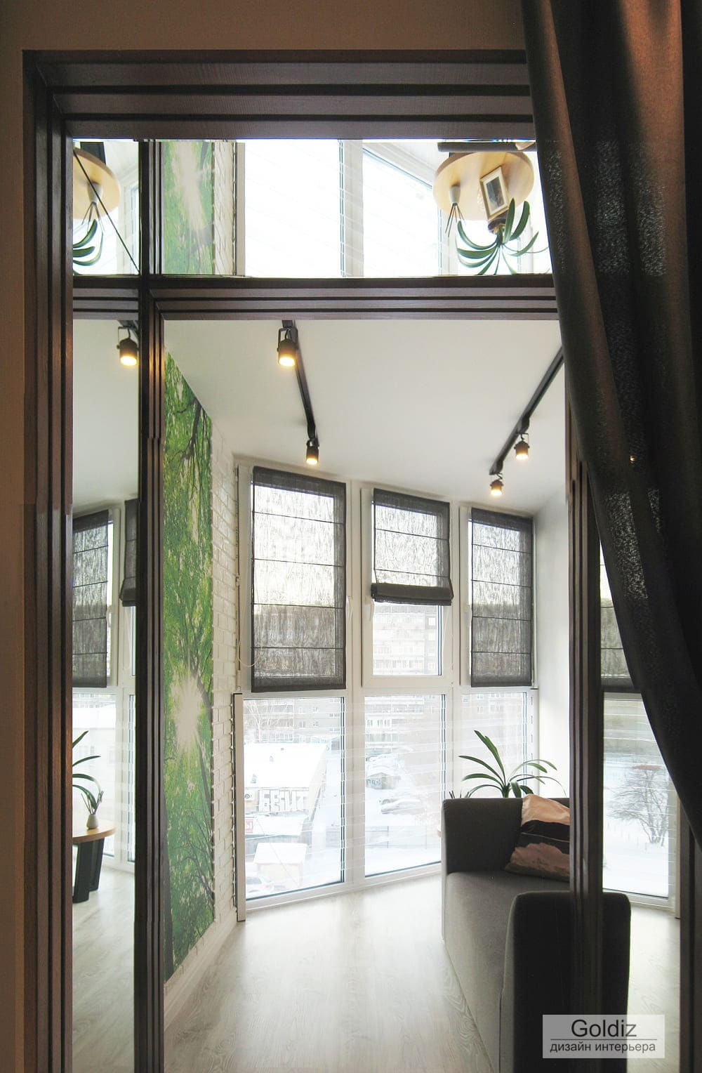 Дизайн лоджии с панорамными окнами, серым диваном и акцентной зелёной фотопанелью, создающей светлое и уютное пространство