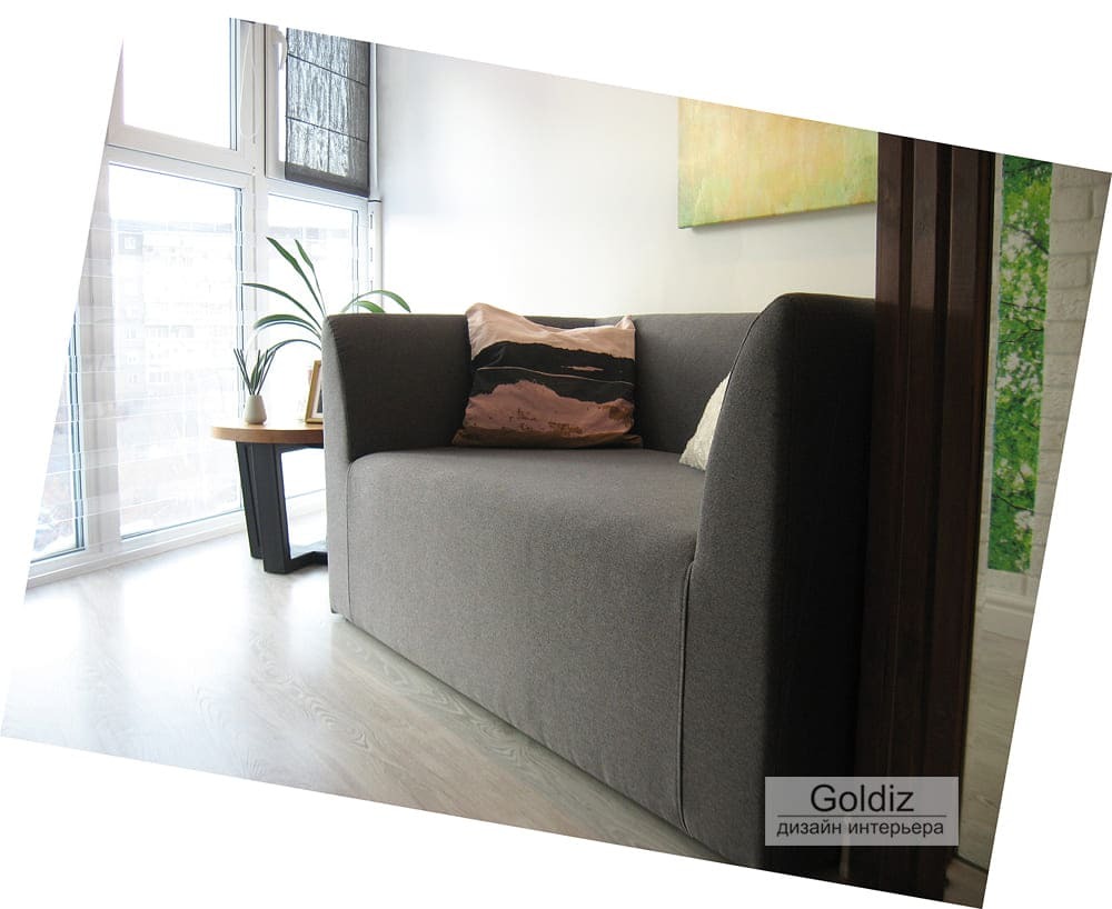 Современный дизайн лоджии с панорамными окнами, серым компактным диваном и минималистичным оформлением пространства для отдыха