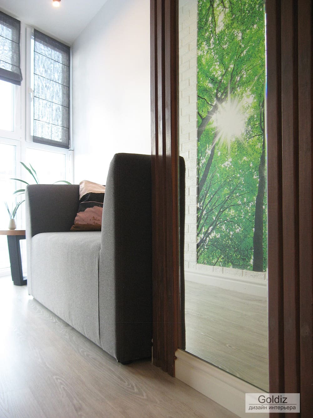 Зона отдыха на лоджии с мягким диваном, римскими шторами, акцентной зелёной панелью и большим остеклением